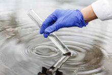 Рука в синей перчатке набирает в прозрачную стеклянную пробирку воду из водоема.